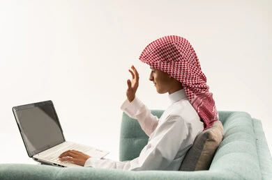 Raising the palm of the hand is a sign of greeting and responding to peace, making a video call using a laptop, exchanging news with family and friends, a side portrait of a young Arab Gulf Saudi man wearing a white thobe and a ghutrah sitting on the sofa communicating with relatives, expressions of joy and happiness, white background.