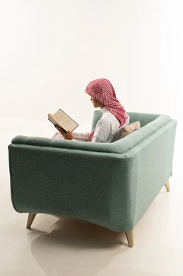 Culture, reading, and exploration, spending free time learning and gaining knowledge, the concept of education and scientific research, a portrait from behind of a young Arab Gulf Saudi man wearing a white thobe and a shemagh sitting on a sofa holding a book, gestures of engagement and focus, white background.