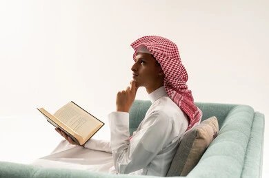 قضاء وقت الفراغ بالتعلم والثقافة، مفهوم التثقيف والبحث العلمي، هواية المطالعة والاستكشاف، بورتريه من الجانب لشاب عربي خليجي سعودي يرتدي الثوب الابيض و الشماغ يجلس على الاريكة يمسك كتاب ويضع يده على ذقنه ينظر إلى شيء ما بتعابير التفكير، خلفية بيضاء