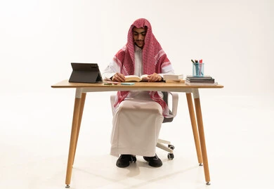 Educational scientific curricula, the concept of diligence and excellence, daily lesson review, school education in the Kingdom of Saudi Arabia, a portrait of an Arab Gulf Saudi student wearing a white thobe and a shemagh sitting at a table in front of a tablet studying school books, white background.