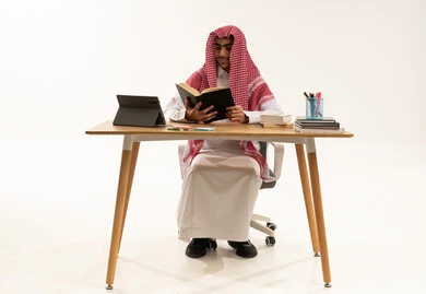 The concept of diligence and excellence, reviewing daily lessons, school education in the Kingdom of Saudi Arabia, a portrait of an Arab Gulf Saudi student wearing a white thobe and a shemagh sitting at a table in front of a tablet studying school books, white background.