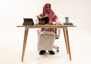 School education in the Kingdom of Saudi Arabia, daily lesson review, the concept of diligence and excellence, a portrait of a Saudi Gulf Arab student wearing a white thobe and a shemagh sitting at a table in front of a tablet studying school books, white background.