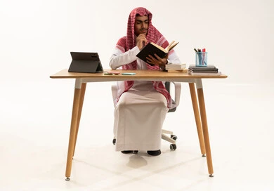 The concept of back to school, educational scientific curricula, a portrait of an Arab Gulf Saudi student wearing a white thobe and a shemagh sitting at a table in front of a tablet, resting his hand on his chin with gestures of questioning and surprise while reading a book, white background.