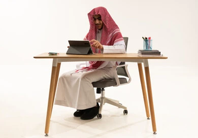 إيماءات التفاعل والمشاركة، متابعة الحصص و حل الواجبات عبر الانترنت ، بورترية لطالب عربي خليجي سعودي يرتدي الثوب الابيض و الشماغ يجلس أمام طاولة الدراسة والتابلت يقوم بمتابعة الدروس عن طريق منصات التعلم عن بعد، خلفية بيضاء
