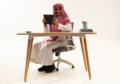 النظر الى الكاميرا بايماءات السعادة و السرور، متابعة الحصص و حل الواجبات عبر الانترنت ، بورترية لطالب عربي خليجي سعودي يرتدي الثوب الابيض و الشماغ يجلس أمام طاولة الدراسة والتابلت يقوم بمتابعة الدروس عن طريق منصات التعلم عن بعد، إيماءات التفاعل والمشاركة، خلفية بيضاء