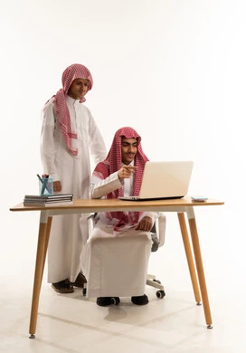 Collaboration in understanding study materials, completing homework, a portrait of a Saudi Gulf Arab student wearing a white thobe and a mischievous expression sitting at a table in front of a laptop sharing information with a classmate, a new classroom, white background.