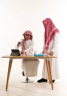 Wearing protective glasses and conducting a chemical experiment, collaboration among students and benefiting from laboratory research, a portrait of an Arab Gulf Saudi student wearing a white thobe and a shemagh sitting at a table in front of a tablet using test tubes to conduct experiments in the lab accompanied by a female colleague, white background.