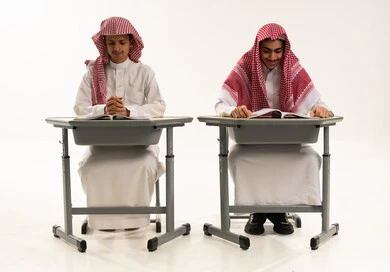 Back to school season, gestures of diligence and excellence, a portrait of two Arab Gulf Saudi students wearing white thobes and shemaghs sitting at a table reviewing lessons for studying and school tests, white background.