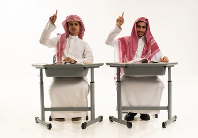 Active participation in lessons, a portrait of two Arab Gulf students from Saudi Arabia wearing white thobes and shemaghs sitting at a table raising their hands to answer, education and teaching with the prescribed curricula for students in the Kingdom of Saudi Arabia, expressions and gestures of enthusiasm to know the correct answer, white background.