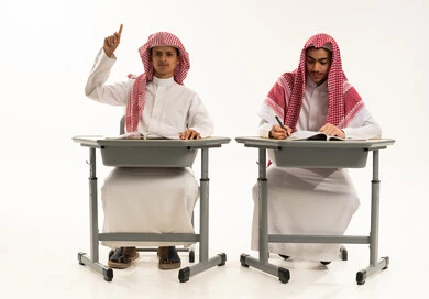 Academic interaction with the teacher and classmates, expressions and gestures of enthusiasm to know the correct answer, completing homework assignments, a portrait of two Arab Gulf Saudi students wearing white thobes and shemaghs sitting at a table in the classroom, education and teaching with the prescribed curricula for students in the Kingdom of Saudi Arabia, white background.