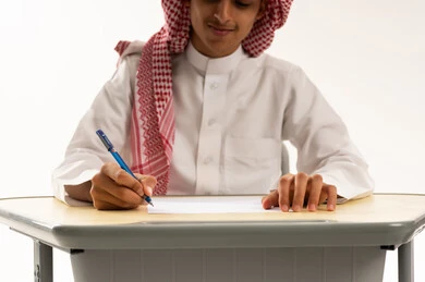 بدء الفصل الدراسي الجديد فى المدارس السعودية, بورترية مقرب لطالب عربي خليجي سعودي يرتدي الثوب الابيض و الشماغ يجلس على الطاولة يقوم بمراجعة الدروس للمذاكرة والاختبارات المدرسية ، خلفية بيضاء