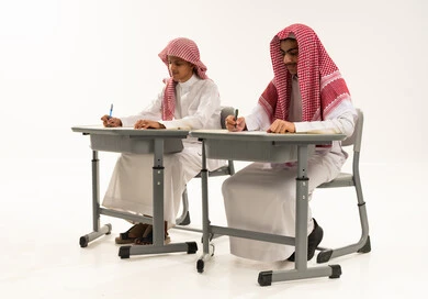 Developing educational curricula in the Arab Kingdom, gestures of diligence and excellence, a portrait of two Arab Gulf students from Saudi Arabia wearing white thobes and shemaghs sitting at a table reviewing lessons for studying and school tests, white background.