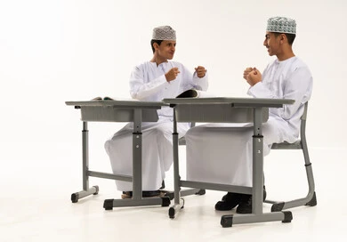 Spending a good time during the school day, a new classroom, a portrait of two Arab Gulf Omani students wearing the dishdasha and kumma sitting at the table enjoying fun and play during the lesson, education in the Sultanate of Oman, white background.