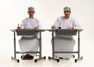 النظر الى الكاميرا بايماءات السعادة و السرور، المذاكرة للإختبارات المدرسية، بورتريه لطالبان عربيان خليجيان عمانيان يرتديان الدشداشة و الكمة يجلسان على الطاولة يقومان بتأدية الواجبات المدرسية، مراجعة الدروس الدراسية، الإستعداد للعام الدراسي الجديد، التعليم الدراسي في سلطنه عمان، العودة إلى المدارس، خلفية بيضاء