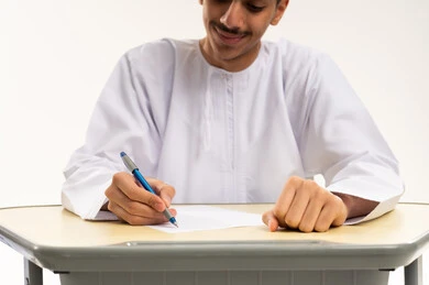 The goals of education, focus and integration during study, the beginning of a new school year, a close portrait of an Arab Gulf Omani student wearing a dishdasha and kumma sitting at a table studying the lesson and diligently writing exercises, the quality of education in schools in the Sultanate of Oman, white background.
