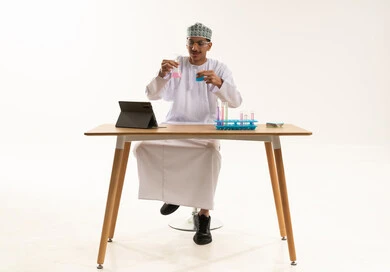 التطوير والبحث وإجراء التفاعلات الكيميائية, الدراسات والتجارب في بيئة عملية متطورة، اكتساب المعرفة والخبرة بإيماءات الدقة والتركيز،  بورتريه لطالب عربي خليجي عماني يرتدي الدشداشة و الكمة يضع نظارة الوقاية يجلس على الطاولة امام جهاز التابلت يقوم بتجربة علمية, خلفية بيضاء