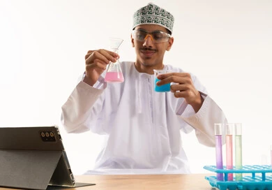 Studies and experiments in an advanced practical environment, acquiring knowledge and experience with gestures of precision and focus, development and research, and conducting chemical reactions. A portrait of an Arab Gulf Omani student wearing a dishdasha and kumma, wearing safety glasses, sitting at a table in front of a tablet conducting a scientific experiment, with a white background.
