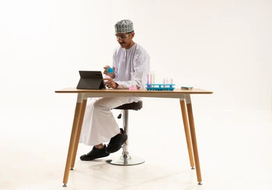 Acquiring knowledge and experience with gestures of precision and focus, studies and experiments in an advanced practical environment, development and research, and conducting chemical reactions. A portrait of an Arab Gulf Omani student wearing a dishdasha and kumma, wearing protective glasses, sitting at a table in front of a tablet conducting a scientific experiment, with a white background.