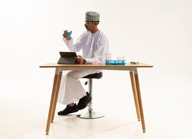 Studies and experiments in an advanced practical environment, acquiring knowledge and experience with gestures of precision and focus, development and research, and conducting chemical reactions. A portrait of an Arab Gulf Omani student wearing a dishdasha and kumma, wearing protective glasses, sitting at a table in front of a tablet conducting a scientific experiment, with a white background.