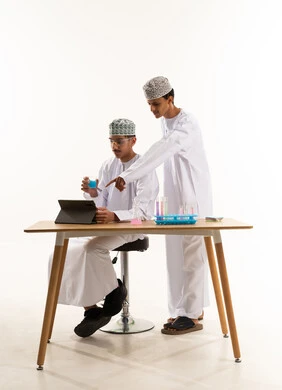 Teamwork and collaboration among colleagues, benefiting from technologies and technological devices in effective learning, using experimental educational tools and means, a portrait of two Arab Gulf Omani students wearing the dishdasha and kumma conducting chemical experiments and tests, white background.
