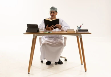 ايماءات الاندماج و التركيز، الاجتهاد وطلب العلا والمثابرة والحرص على النجاح، بورتريه لطالب عربى خليجي عماني يرتدي الدشداشة و الكمه يجلس على الطاولة المكتبيه يقوم بالمذاكرة للاختبارات والاستعداد للامتحان، خلفية بيضاء