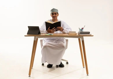 النظر الى  الكاميرا بايماءات السعادة و السرور، الاجتهاد وطلب العلا والمثابرة والحرص على النجاح، بورتريه لطالب عربى خليجي عماني يرتدي الدشداشة و الكمه يجلس على الطاولة المكتبيه يقوم بالمذاكرة للاختبارات والاستعداد للامتحان، خلفية بيضاء