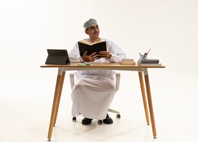 النظر الى شيء ما، الاجتهاد وطلب العلا والمثابرة والحرص على النجاح، بورتريه لطالب عربى خليجي عماني يرتدي الدشداشة و الكمه يجلس على الطاولة المكتبيه يقوم بالمذاكرة للاختبارات والاستعداد للامتحان، خلفية بيضاء