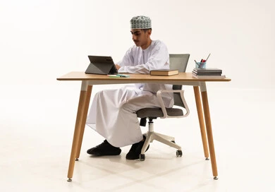 The development of teaching methods in schools, benefiting from modern technologies and technology in learning and studying, developed curricula and e-learning, a portrait of an Arab Gulf Omani student wearing a dishdasha and kumma sitting at a table using a tablet, white background.