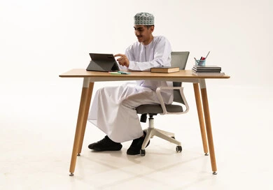 الاستفادة من التقنيات والتكنولوجيا الحديثة في التعلم والدراسة، المناهج المطورة والتعليم الالكتروني، تطور طرائق التدريس في المدارس ، بورتريه لطالب عربي خليجي عماني يرتدي الدشداشة و الكمة يجلس على الطاولة يستخدم جهاز التابلت، خلفية بيضاء