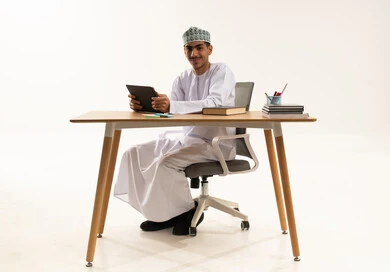 الإستعداد للمحاضرات الالكترونية ،  النظر الى الكاميرا بايماءات السعادة و السرور، بورتريه لطالب عربي خليجي عماني يرتدي الدشداشة و الكمة يجلس على الطاولة يستخدم جهاز التابلت ، التعليم في سلطنه عمان، خلفية بيضاء