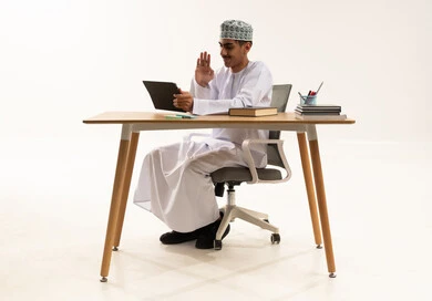 دمج الأجهزة التقنية الحديثة بالتعلم،الاستعداد للعام الدراسي الجديد, مفهوم العودة إلى المدارس، رفع اليد بتعابير القاء التحية والسلام، بورتريه لطالب عربي خليجي عماني يرتدي الدشداشة و الكمة يجلس على الطاولة يستخدم جهاز التابلت و يقوم باجراء مكالمة فيديو ، خلفية بيضاء