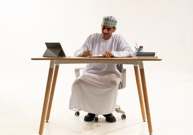 Focus and engagement during study, the beginning of a new school year, the goals of education, a portrait of an Arab Gulf Omani student wearing a dishdasha and kumma sitting at a table studying the lesson and diligently writing exercises, the quality of education in schools in the Sultanate of Oman, white background.