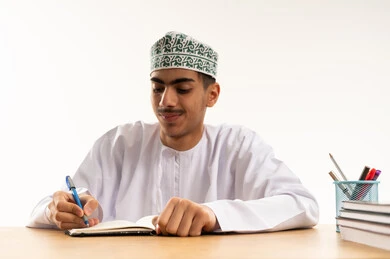 الاجتهاد للوصول إلى النجاح والتفوق، موسم العودة للمدارس، بورتريه لطالب عربي خليجي عماني يرتدي الدشداشة و الكمة يجلس على الطاولة يقوم بحل الواجبات والمراجعة، خلفية بيضاء