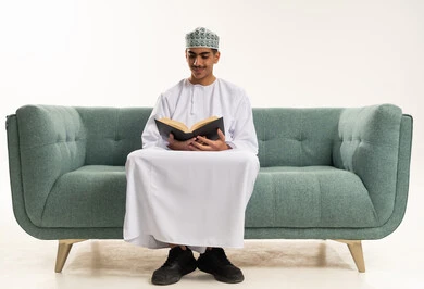 قراءة الكتب بتمعن، ثقافة التعلم الذاتي ،  بورترية لشاب عربي خليجي عماني يرتدي الدشداشة و الكمة يجلس على الاريكة يحمل بيده كتاب تثقيفي ، الاستمتاع بقراءة الروايات والقصص، خلفية بيضاء