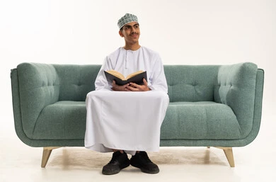 Looking at something, the culture of self-learning, reading books attentively, a portrait of a young Arab Gulf Omani man wearing a dishdasha and kumma sitting on a sofa holding an educational book, enjoying reading novels and stories, white background.