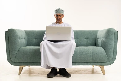 إجراء مكالمة فيديو، رفع كف اليد بايماءات القاء التحية والسلام، التعلم عن بعد وتلقي الدروس عبر الأونلاين، أنشطة ترفيهية الكترونية مسلية، استخدام التكنولوجيا والتقنية الحديثة والمتطورة، بورتريه لشاب عربي خليجي عماني يرتدي الدشداشة و الكمة يجلس على الاريكة يستخدم جهاز الحاسوب المحمول ، خلفية بيضاء