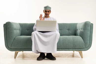 Raising the hand with gestures of greeting and peace, making a video call, distance learning and receiving lessons online, entertaining electronic recreational activities, using modern and advanced technology and techniques, a portrait of a young Arab Gulf Omani man wearing a dishdasha and kumma sitting on a sofa using a laptop, white background.