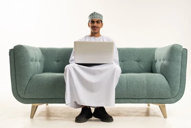 Distance learning and receiving lessons online, entertaining electronic recreational activities, using modern and advanced technology, spending enjoyable times, a portrait of a young Arab Gulf Omani man wearing a dishdasha and kumma sitting on a sofa using a laptop, white background.