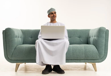 Fun electronic recreational activities, distance learning and taking lessons online, using modern and advanced technology, spending enjoyable times, a portrait of a young Arab Gulf Omani man wearing a dishdasha and kumma sitting on a sofa using a laptop and looking at something, white background.