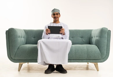 استخدام التكنولوجيا والتقنيات الحديثة, تصفح مواقع التواصل الاجتماعي، متابعة الأفلام والمسلسلات الممتعة، المحادثة مع الأهل والأصدقاء، بورتريه لشاب عربي خليجي عماني يرتدي الدشداشة و الكمة يجلس على الاريكة يحمل بيده جهاز التابلت, ايماءات السعادة و السرور، خلفية بيضاء