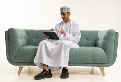 متابعه آخر أخبار السوشال ميديا، بورتريه لشاب عربي خليجي عماني يرتدي الدشداشة و الكمة يجلس على الاريكة يحمل بيده جهاز التابليت، التسويق الإلكتروني عبر الأجهزة الحديثة، تصفح مواقع التواصل الإجتماعي، خلفية بيضاء