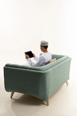 التواصل و الدردشة مع الاقارب، استخدام تطبيقات وبرامج الأجهزة اللوحية, التطور التكنولوجي والتقني في مجالات العمل عن بعد، بورتريه من الخلف لشاب عربي خليجي عماني يرتدي الدشداشة و الكمة يجلس على الاريكة يحمل بيده جهاز التابلت، تصفح مواقع التواصل الاجتماعي، خلفية بيضاء