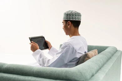 استخدام تطبيقات وبرامج الأجهزة اللوحية، التواصل و الدردشة مع الاقارب,التطور التكنولوجي والتقني في مجالات العمل عن بعد، بورتريه من الخلف لشاب عربي خليجي عماني يرتدي الدشداشة و الكمة يجلس على الاريكة يحمل بيده جهاز التابلت، تصفح مواقع التواصل الاجتماعي، خلفية بيضاء