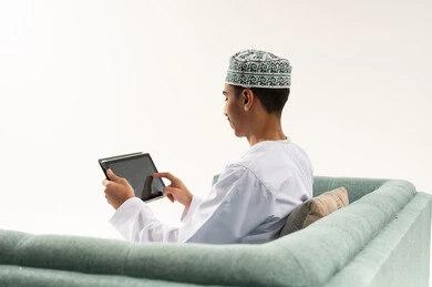 التواصل و الدردشة مع الاقارب,التطور التكنولوجي والتقني في مجالات العمل عن بعد، بورتريه من الخلف لشاب عربي خليجي عماني يرتدي الدشداشة و الكمة يجلس على الاريكة يحمل بيده جهاز التابلت، تصفح مواقع التواصل الاجتماعي، استخدام تطبيقات وبرامج الأجهزة اللوحية، خلفية بيضاء