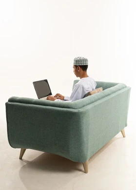 Typing on a keyboard, browsing social media, using a laptop to complete tasks, a portrait from behind of a young Arab Gulf Omani man wearing a dishdasha and kumma sitting on a sofa holding a laptop and making a video call, the evolution of technology and technical devices, white background.