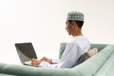تصفح برامج التواصل الاجتماعي, إستخدام الحاسوب المحمول لإنجاز المهام، بورتريه من الخلف لشاب عربي خليجي عماني يرتدي الدشداشة و الكمة يجلس على الاريكة يحمل جهاز اللابتوب و يقوم باجراء مكالمة فيديو ، تطور التكنولوجيا والأجهزة التقنية، خلفية بيضاء