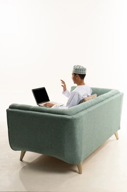 تصفح برامج التواصل الاجتماعي، رفع اليد بتعابير القاء التحية والسلام, إستخدام الحاسوب المحمول لإنجاز المهام، بورتريه من الخلف لشاب عربي خليجي عماني يرتدي الدشداشة و الكمة يجلس على الاريكة يحمل جهاز اللابتوب و يجري مكالمة فيديو ، تطور التكنولوجيا والأجهزة التقنية، خلفية بيضاء