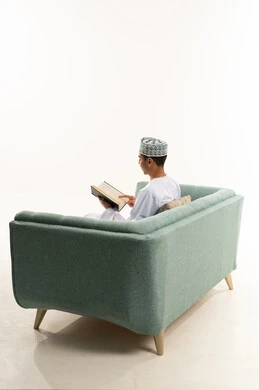 التسلية بقراءة الروايات والقصص، الأنشطة الترفيهية المفيدة في وقت الفراغ، مفهوم الثقافة والتعلم الذاتي، ممارسة هواية المطالعة, بورتريه من الخلف لشاب عربي خليجي عماني يرتدي الدشداشه و الكمه يجلس على الاريكة يقوم بقراءة كتاب, خلفية بيضاء