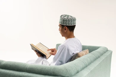 الاستمتاع بقراءة الروايات الأدبية، الأنشطة الترفيهية المفيدة في وقت الفراغ، مفهوم الثقافة والتعلم الذاتي، ممارسة هواية المطالعة, بورتريه من الخلف لشاب عربي خليجي عماني يرتدي الدشداشه و الكمه يجلس على الاريكة يقوم بقراءة كتاب, خلفية بيضاء