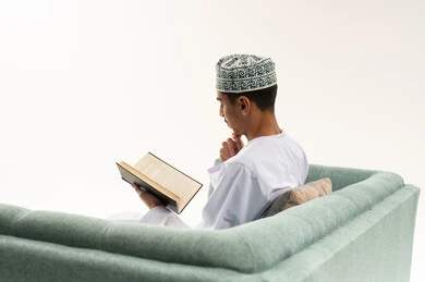 الاستمتاع بالأنشطة الترفيهية المفيدة في وقت الفراغ, قراءة القصص والروايات، مفهوم الثقافة والتعلم الذاتي، ممارسة هواية المطالعة, بورتريه من الخلف لشاب عربي خليجي عماني يرتدي الدشداشه و الكمه يجلس على الاريكة يحمل بيده كتاب ينظر بايماءات التفكير, خلفية بيضاء